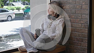 Sitting on windowsill blonde woman enjoys hot coffee looks thoughtfully through window. Warm hot coffee contrasts with