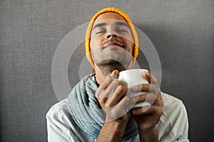 Sitting young handsome man with a mug of coffee, tea, water. Dreaming and smiling with closed eyes on gray background.