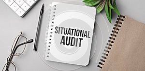 situational audit is written in a white notebook with calculator, craft colored notepad, plant, black marker and glasses