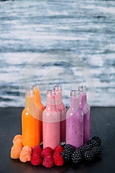 Six bottles with smoothies and raspberry, red, yellow, blackberry on black table grey background. Milk shake in glass jar