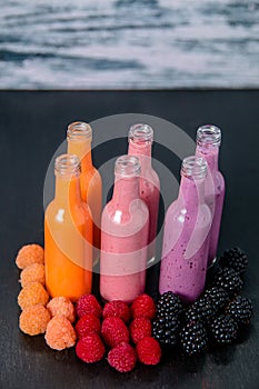 Six bottles with smoothies and raspberry, red, yellow, blackberry on black table grey background. Milk shake in glass jar