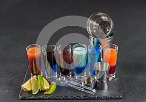 six different colored shot drinks, lined up on a black stone plate, ice cubes in shaker and ice tongs, lemon and lime