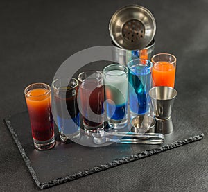 six different colored shot drinks, lined up on a black stone plate, ice cubes in shaker and ice tongs