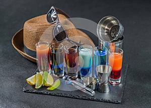 six different colored shot drinks, lined up on a black stone plate, ice cubes in shaker and ice tongs, lemon and lime, brown