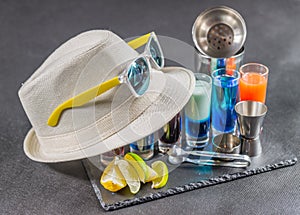 six different colored shot drinks, lined up on a black stone plate, ice cubes in shaker and ice tongs, lemon and lime, white