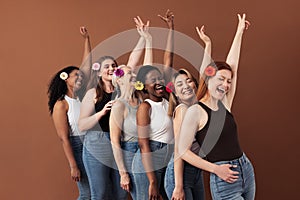 Six laughing women with flowers in hair. Multi-ethnic group of females raises hands up over brown background