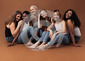 Six women of different ages sitting together in studio on brown background. Multi-ethnic group of diverse females having good