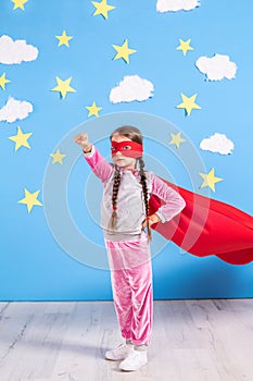 Six year blonde girl dressed like superhero having fun at home. Kid on the background of bright blue wall.