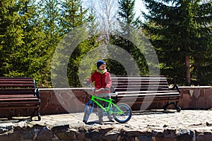 Six-year-old boy in a warm jacket and hat holds a two-wheeled bike outdoors, wooden benches