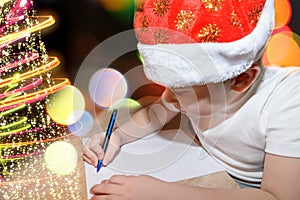 Six year old cute boy with a red christmas cap writing a letter to Santa Claus near the Christmas tree indoors. Bright New Year ba