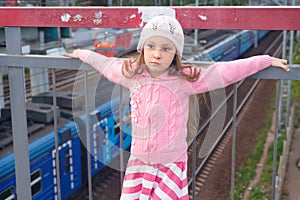 a six-year-old girl in a pink coil stands on a bridge over the railroad tracks. a passing train is visible