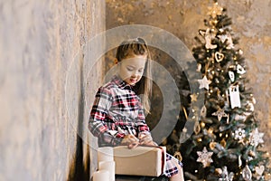 Six-year-old girl in a plaid dress on the background of a Christmas tree opens her new year gift