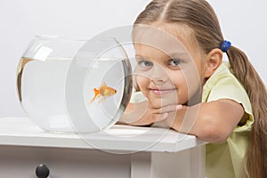 Six year old girl put her head on the handle sitting at the table with an aquarium with goldfish