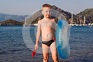 A six year old tanned boy stands holding a swimming circle and a sand shovel by the sea