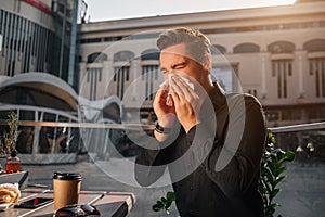 Sixk young man sit at table and sneezing in white napkin. He keeps eyes closed. Guy suffer. He sits outside. It is sunny.