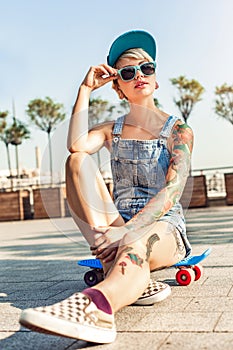 Skateboarding. Alternative girl skater in cap and sunglasses sitting on penny board on the city street looking aside
