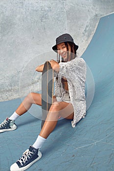 Skateboarding. Asian Girl In Urban Outfit With Skateboard Sitting On Ramp Against Concrete Wall At Skatepark.