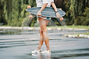 Skater girl standing crossed legs holding her longboard behind h