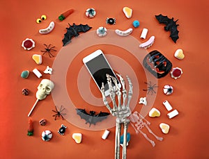 Skeleton using smartphone. Halloween decorations on orange background