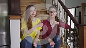 Skeptic young woman in eyeglasses showing thumb down as cheerful twin sister gesturing thumb up smiling looking at