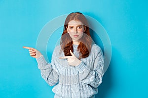 Skeptical and displeased redhead girl pointing fingers left with upset face, standing disappointed against blue