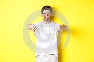 Skeptical guy showing thumbs down and smirking, dislike and disagree, standing over yellow background