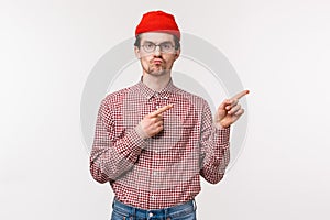 Skeptical serious-looking bothered young geeky man in red beanie and glasses, pouting with unsatisfied grimace pointing