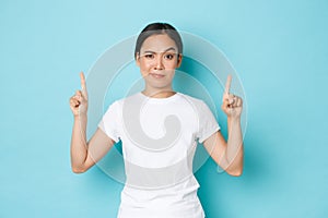 Skeptical and suspicious asian female in white t-shirt, frowning dubious, pointing fingers up at empty space, waiting