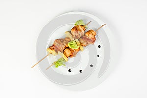 Skewers of chicken and pineapple with bacon on a round white plate on white isolated background