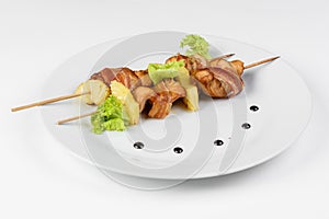 Skewers of chicken and pineapple with bacon on a round white plate on white isolated background