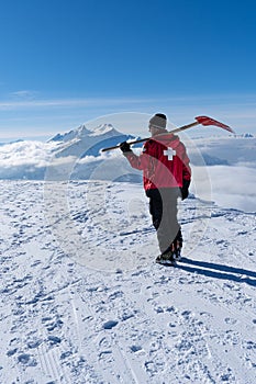 Ski patroller on snow caped mountain is walking against sun with a red rescue jacket and a shovel on his shoulders