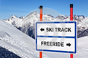 Ski track and freeride delimitation sign with arrows showing opposite directions against snowy mountain  blue sky winter