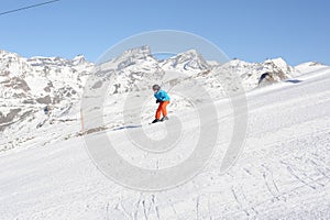 Skier in blue jacket, black helmet and orange pants on the piste slope in winter with snow mountains in Alps