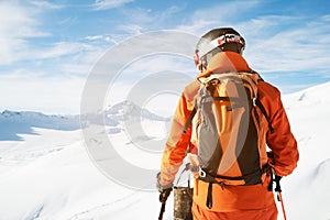 A skier in an orange overall with a backpack on his back wearing a helmet and with ski poles in his hands is standing on