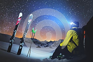 A skier sits at a stone in the mountains at night against a starry sky next to skis and sticks. The concept of extreme
