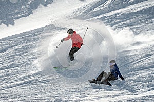 a skier sliding down a snow-covered slope next to a seated snowboarder