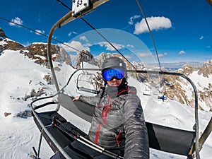 Skier wearing a helmet sitting in ski lift cabin taking a selfie with a camera on monopod.