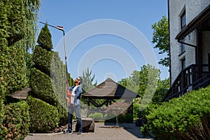 Skilful male landscaper using high altitude hedge trimmer for topiary, shaping thuja in park.