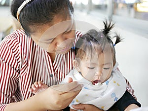 Skilled Asian mother using small spoon to feed her baby girl, 12 months old, at a cafeteria