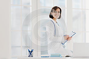 Skilled female physician writes in clipboard, poses at workplace with laptop computer. Professional family doctor ready to give