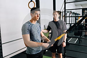 Skilled fitness instructor male showing suspension training to motivated beginner young man, performing pull-ups