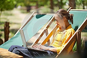 Skilled freelance woman using laptop computer while sitting on sun lounger outdoors, online working digital