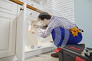 Skilled male worker plumbing repair service installs siphon under sink in kitchen in apartment.