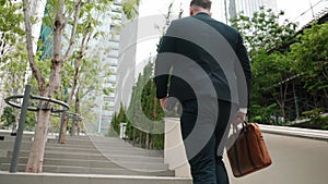 Skilled manager walking up stairs while holding suitcase in green city. Urbane..