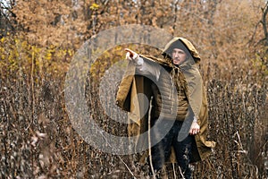 Skilled survivalist male wearing green raincoat tent standing in thicket of bushes in cold overcast day and pointing photo