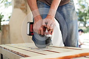 Skilled young male worker is using power screwdriver drilling during construction wooden bench, do it yourself