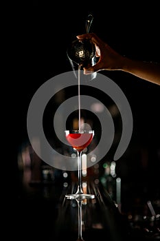 Skillfull bartender woman pours thin stream of alcoholic cocktail into drinking glass standing on the bar counter