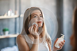Skin care. Beautiful millennial woman removing makeup from her face near mirror at home