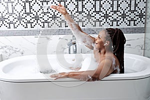 Skin care, clean body, at home during covid. Calm african black lady in bathtub with foam, play splashing with water