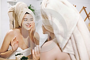 Skin Care concept. Young happy woman in towel making facial massage with organic face scrub and looking at mirror in stylish